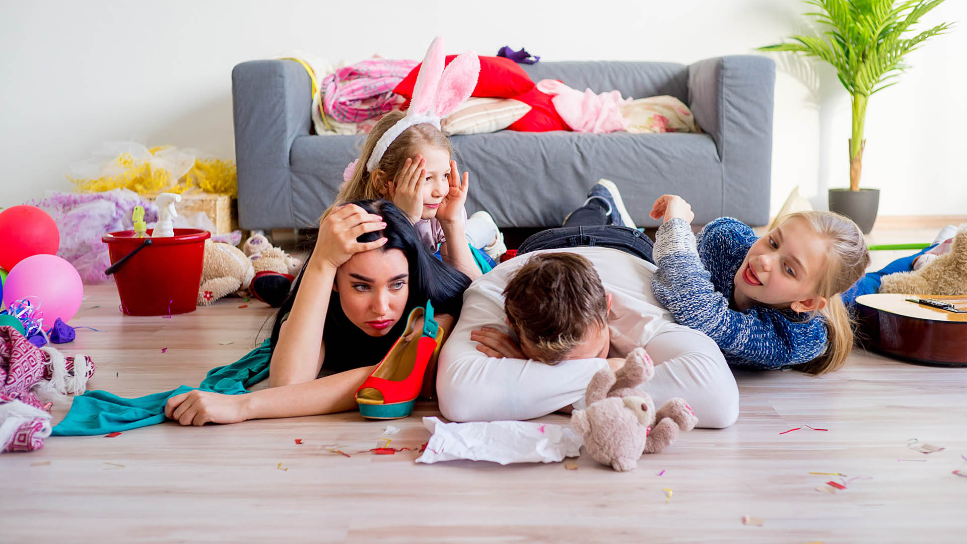 Photo of frustrated parents on the floor of a messy living room with kids hanging all over them